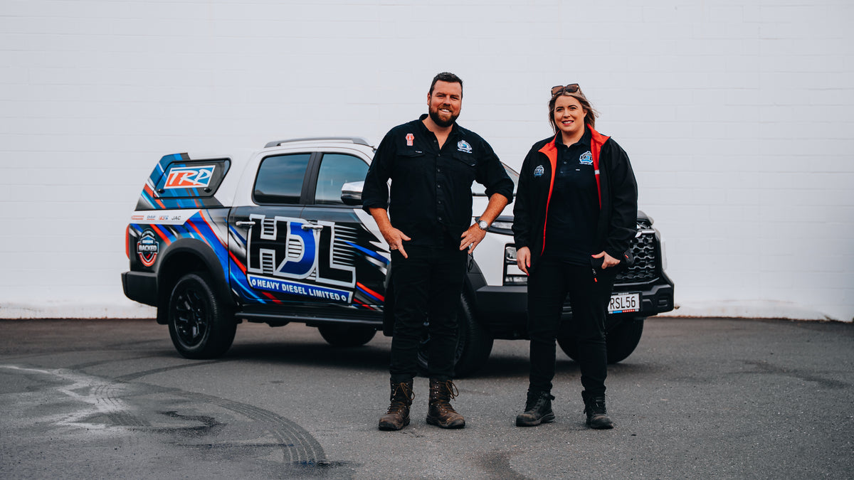 Two people in work uniforms stand smiling in front of a branded pickup truck with “HDL” and colorful graphics on the side, parked on a paved surface with a white wall in the background.