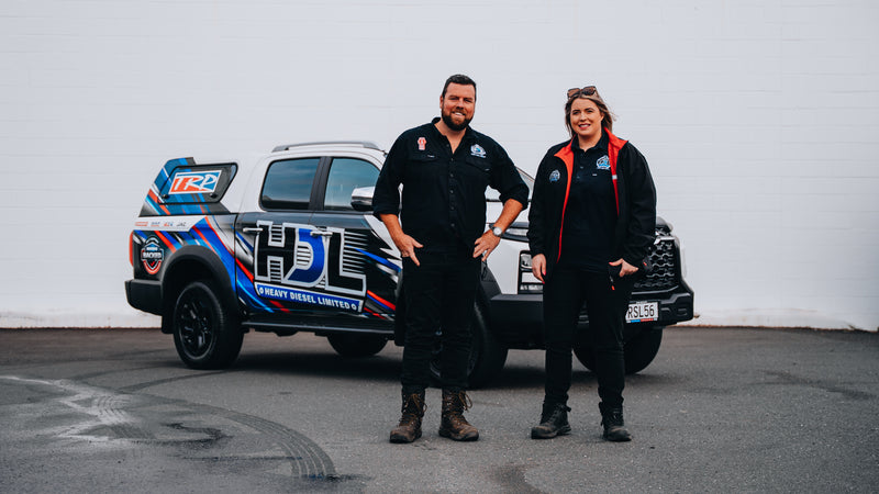 Two people in work uniforms stand smiling in front of a branded pickup truck with “HDL” and colorful graphics on the side, parked on a paved surface with a white wall in the background.