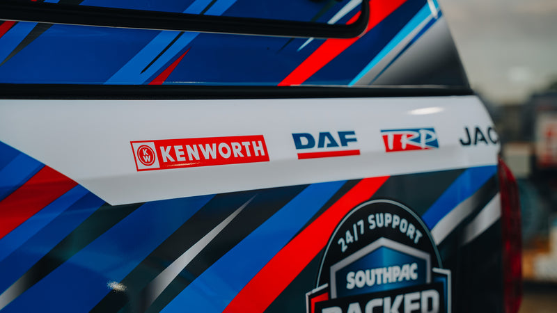 Close-up of a vehicle with colorful blue, red, and white stripes, displaying the logos of Kenworth, DAF, and TRP, along with a 24/7 Support Southpac badge on the side.