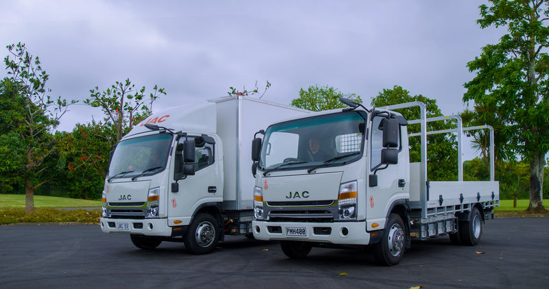 Two white JAC trucks are parked on an asphalt surface, with trees and greenery in the background under a cloudy sky. One truck has a closed box body, while the other has an open flatbed.