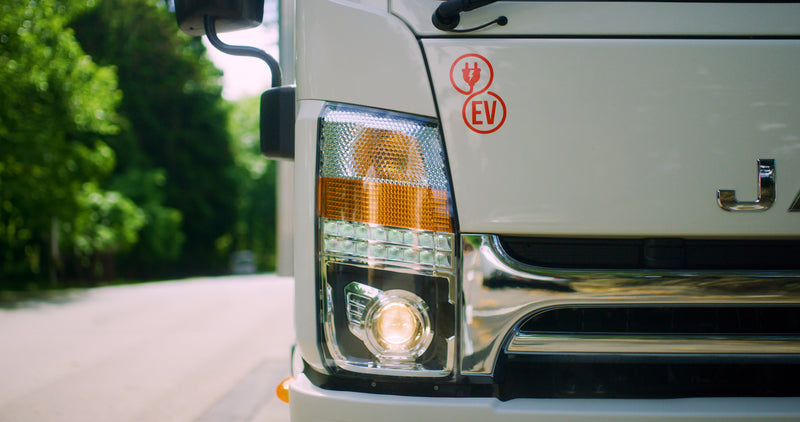 Close-up of the front left side of a white electric vehicle truck with its headlight on. A red EV (electric vehicle) plug icon is visible above the headlight. Trees line the road in the background.