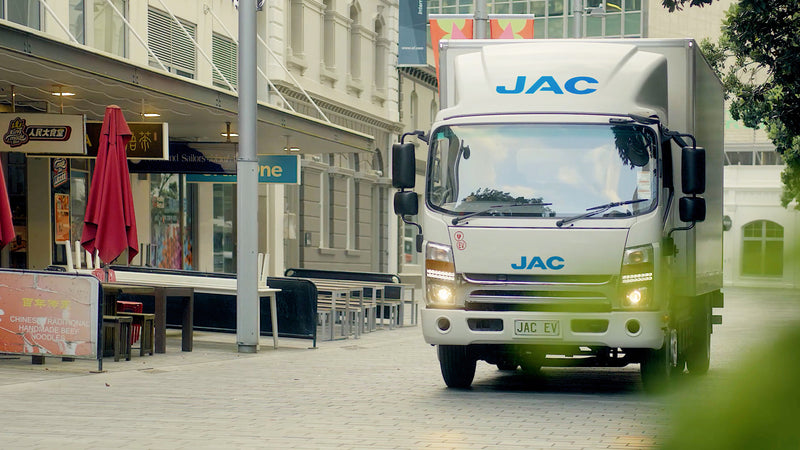 A white JAC delivery truck drives down a pedestrian street lined with restaurants and outdoor seating; red umbrellas and signs are visible on the left.