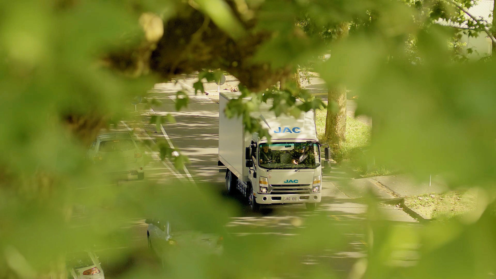 A white JAC delivery truck drives down a sunlit, tree-lined street, partially obscured by green leaves in the foreground.