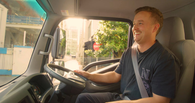A smiling man wearing a seatbelt drives a vehicle through a sunny city street, with sunlight streaming through the window and buildings visible outside.
