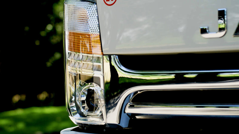 Close-up of the front right headlight and grille of a white vehicle, featuring reflective chrome details and part of the vehicle’s name visible on the hood. Green, blurred background.