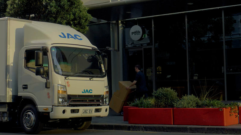 A white JAC delivery truck is parked on the street while a person carries a large cardboard box toward the entrance of a modern building with glass doors and red planters outside.