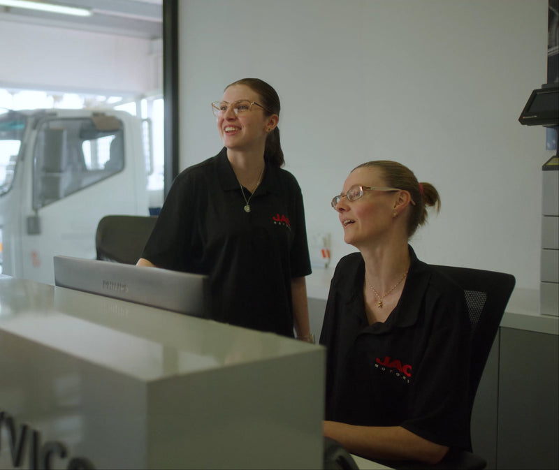 Two women wearing black shirts with red logos are sitting and standing at a reception desk, smiling and looking at a computer monitor in a modern office setting. A truck is visible through the window behind them.