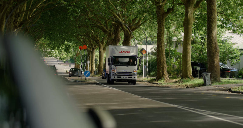 A white delivery truck labeled JAC drives down a tree-lined street in daylight, with sunlight filtering through the green leaves. Other vehicles and street signs are visible along the road.