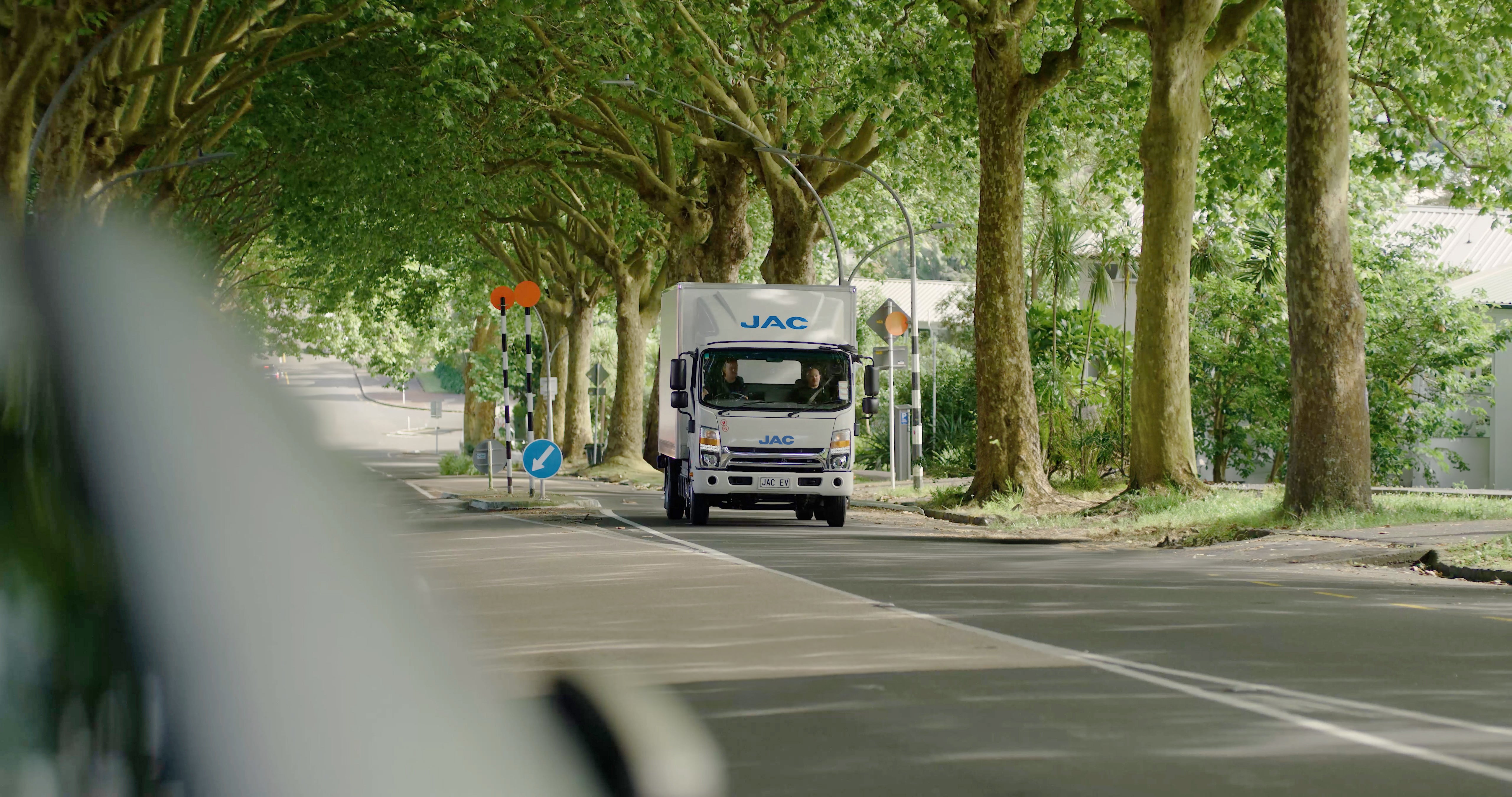 A white JAC delivery truck drives down a tree-lined street with large green trees on both sides, casting shade over the road. Road signs and a fence are visible in the background. The truck is centered in the image.