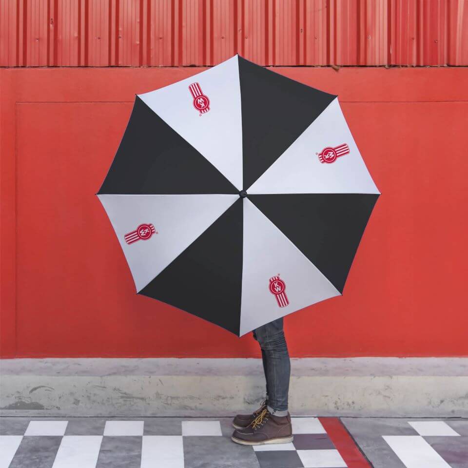 A person stands against a red wall holding the UMBRELLA KW THE BEAST, which obscures their upper body. Only their legs in jeans and brown boots are visible on the checkered pavement.