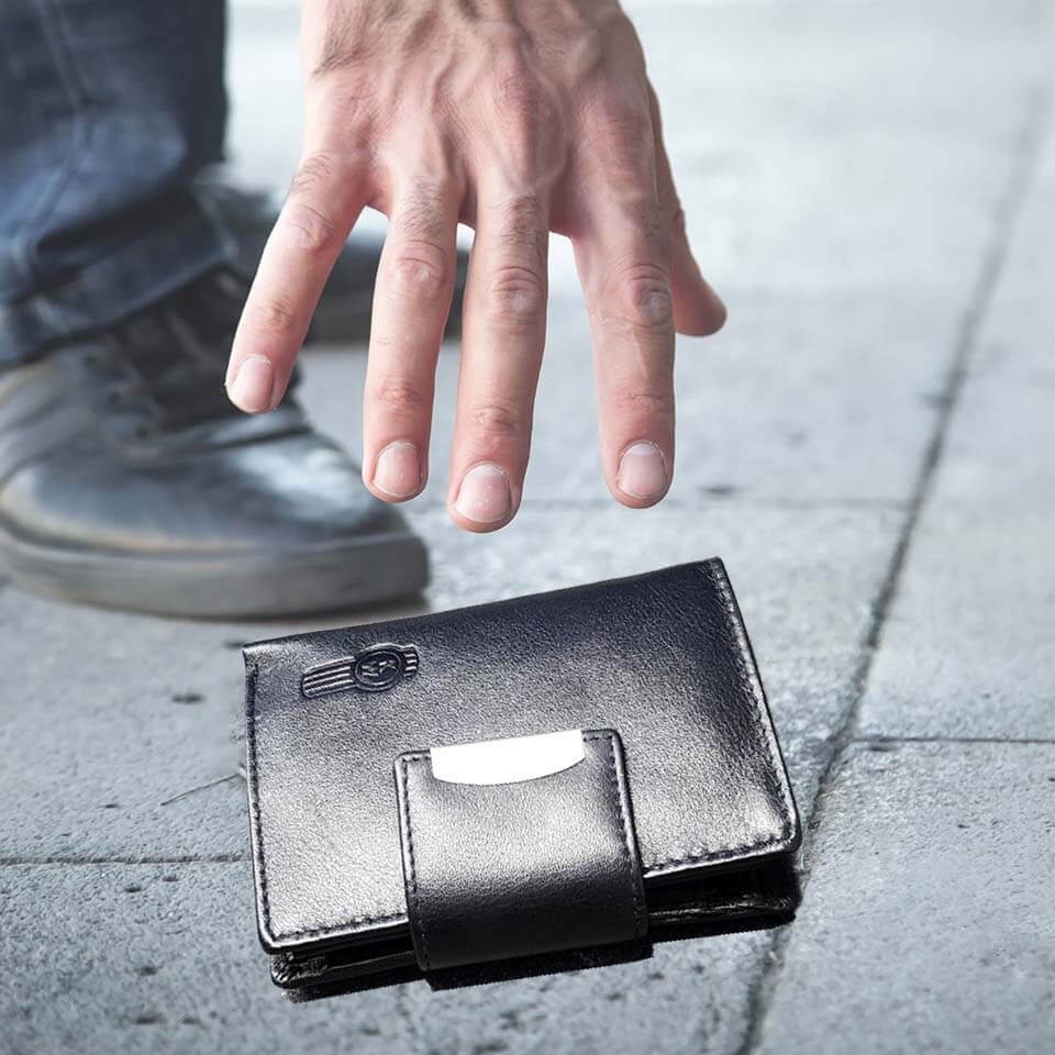 A hand picks up the WALLET KW LEATHER BLACK from a gray sidewalk, showing its smart storage, with a shoe visible in the background.