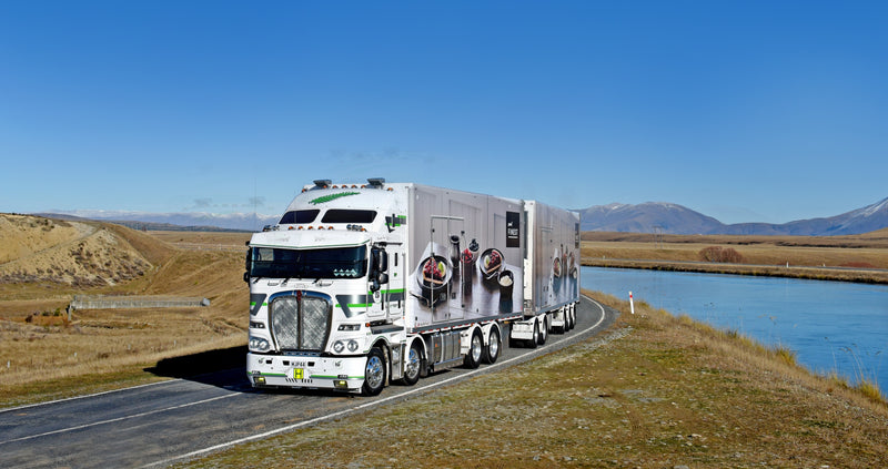 A white semi-truck with a decorated trailer drives along a rural road beside a canal, with grassy hills and distant mountains under a clear blue sky.