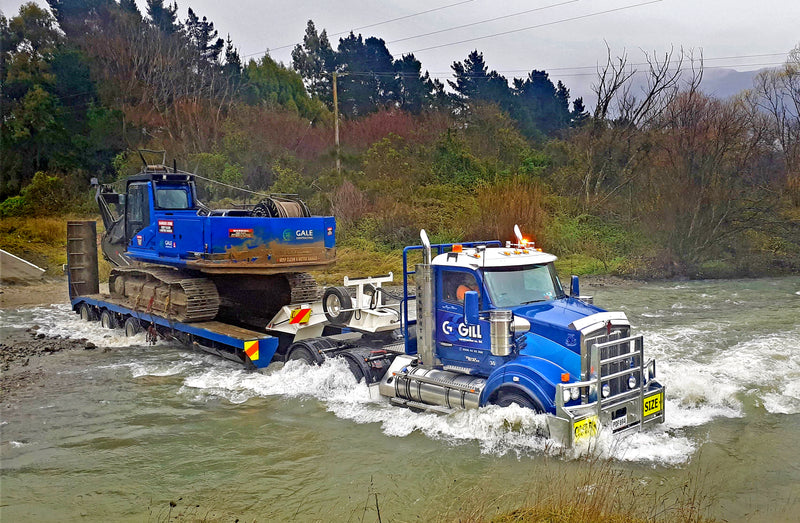A blue semi-truck carrying a blue excavator on a trailer drives through a shallow, flowing river, surrounded by trees and cloudy skies.