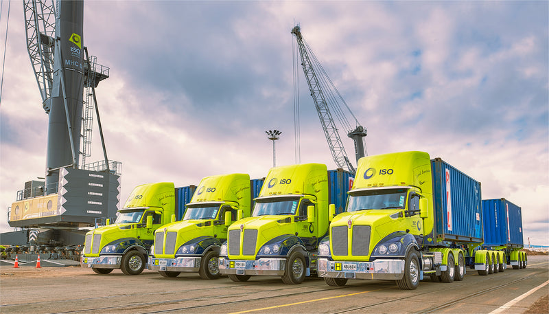 A row of bright yellow-green ISO-branded semi-trucks with blue shipping containers are lined up at a port, with a large crane and cloudy sky in the background.