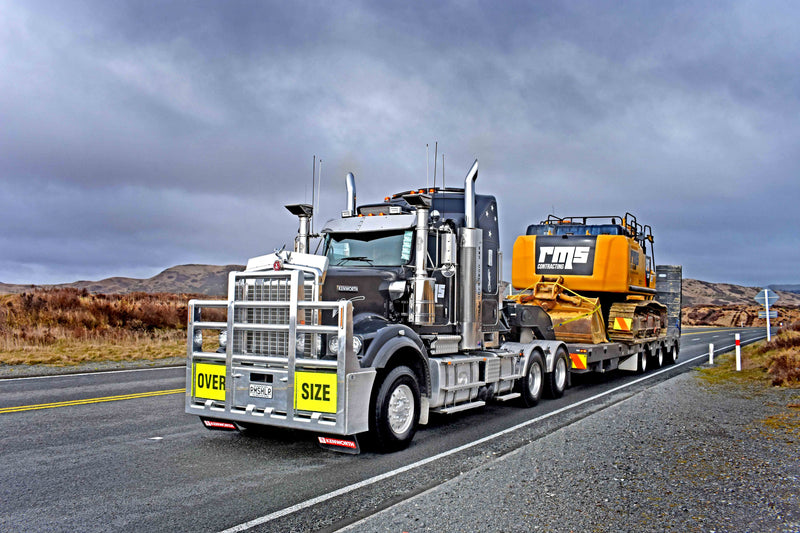 A large semi-truck with OVER SIZE signs hauls a yellow construction excavator on a flatbed trailer along an empty rural road, with cloudy skies and hills in the background.