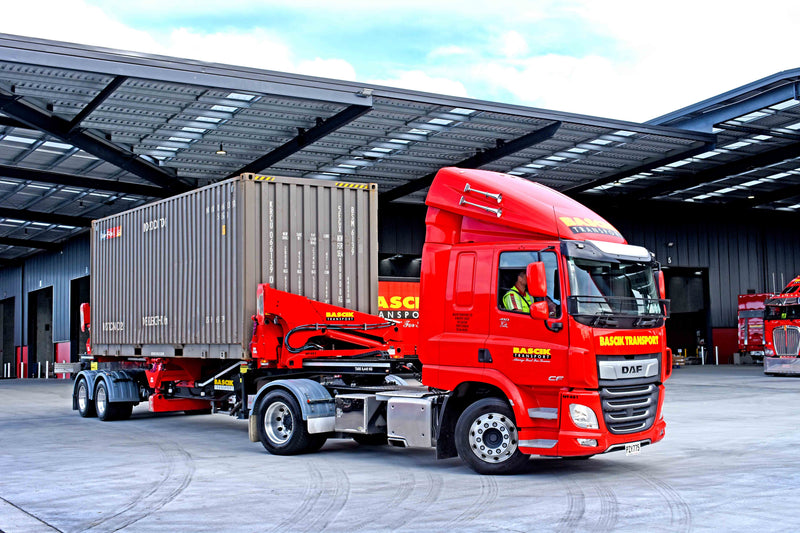 A red Mack Transport truck with a mounted crane is hauling a large shipping container on a trailer outside a warehouse. The driver is visible through the window, and another truck is parked in the background.