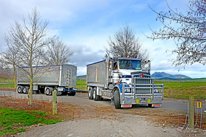 A large silver semi-truck with a trailer is driving on a rural dirt road surrounded by bare trees and green fields, with mountains visible in the background under a partly cloudy sky.