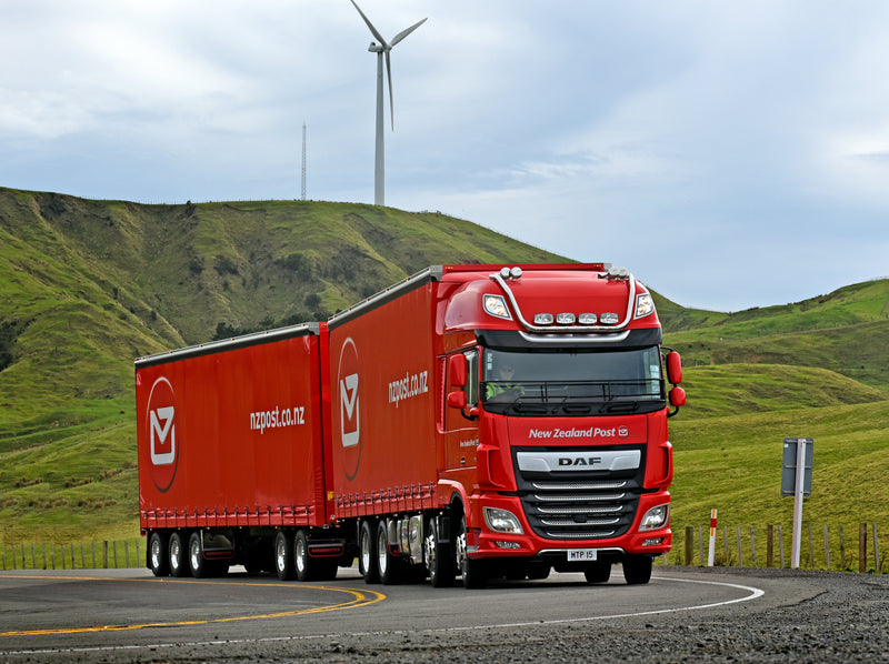 A red New Zealand Post truck with trailers drives on a curved road through green hills, with a wind turbine and cloudy sky in the background.
