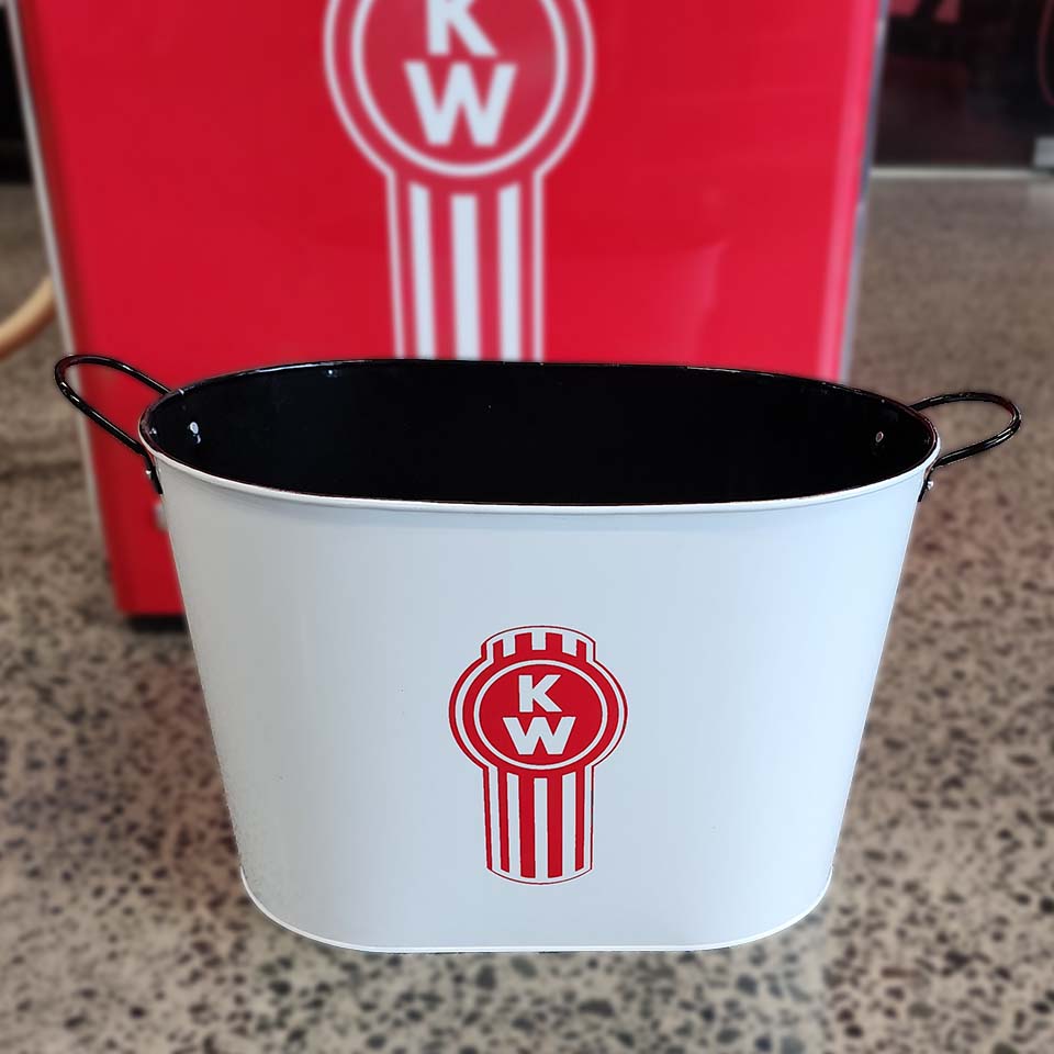 The BUCKET KW ICE/EVENT is a white Kenworth ice tub with black handles and a red Kenworth logo on the front, perfect for BBQs to keep drinks cold. It sits on a speckled floor with a larger matching logo shown in the background.