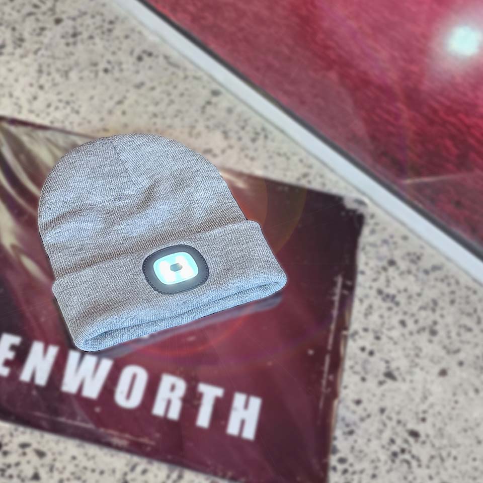A black LED beanie, the BEANIE KW BLACK LED LIGHT, lies on a textured surface, partly over a maroon "ENWORTH" sign—combining practicality and style just like a classic Kenworth hat.