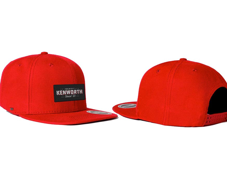 Two vivid CAP KW HAULIN 24-7 RED hats are shown: one displays the structured crown with a black rectangular Kenworth Since 23 patch, while the other features the easily adjustable snapback strap at the back.