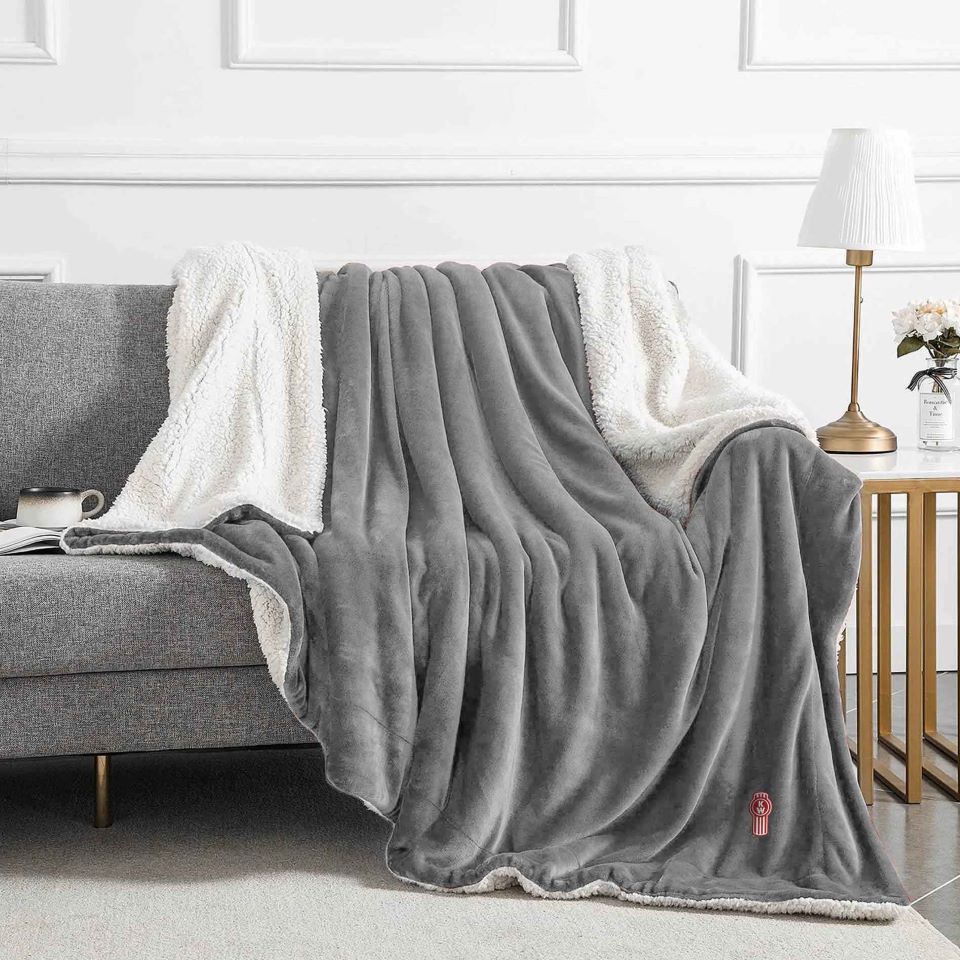 The BLANKET KW GREY/WHIT FLEECE, shown on a modern light gray sofa with a lamp, flowers, and cup nearby, is perfect as a travel blanket or paired with a reusable carry bag for easy storage.