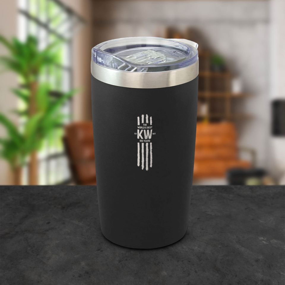 The MUG KW TRAVEL BLACK S/S with double-wall insulation, clear lid, and white KW logo rests on a dark counter. This 300ml stainless steel mug is shown in a modern living room with plants and shelves in the background.
