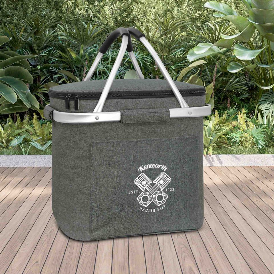 The COOLER KW BASKET 16L, a gray insulated picnic basket with superior insulation and aluminum handles, features a Kenworth logo and engine graphic on the front. It sits on a wooden deck surrounded by lush green plants.