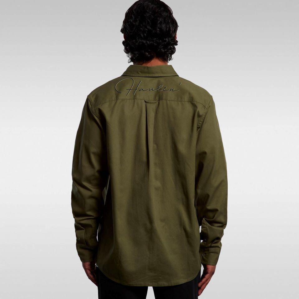 A person faces away, wearing the SHIRT KW DRILL ARMY—an olive green smart long sleeve shirt with "Hawkers" cursive text on the upper back. The background is plain, light gray.