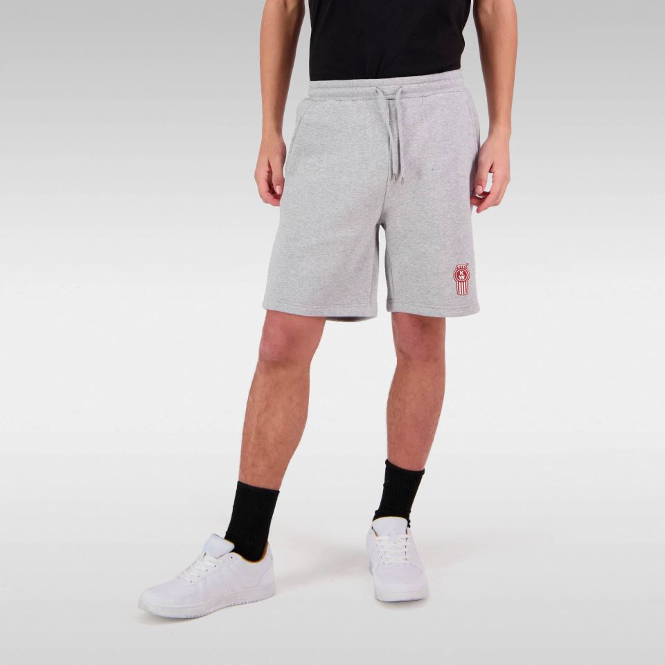 A person wears a black t-shirt, TRACK SHORT KW GREY men's shorts with an embroidered patch, black socks, and white sneakers while standing against a light gray background. Their face is not visible.
