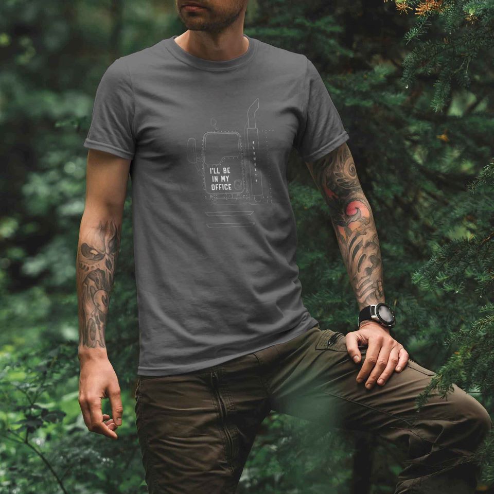 A tattooed person stands in a forest, wearing the TEE SHIRT KW IN MY OFFICE—a comfortable shirt with a truck graphic and "ILL BE IN MY OFFICE"—while resting one knee on a surface and looking to the side.