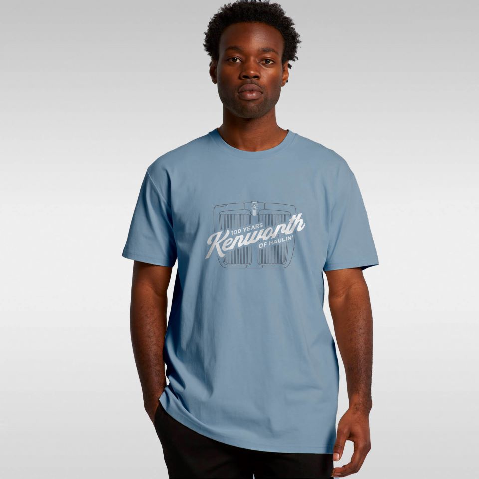 A man wearing a TEE SHIRT KW HAULIN BLUE, featuring a heritage "100 Years Kenworth of Hayward" design, stands against a plain light gray background.