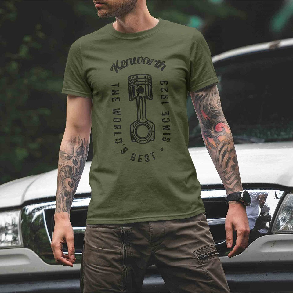 A tattooed man wears a TEE SHIRT KW WORLDS GREEN featuring "The World's Best Since 1923" and a piston graphic while standing in front of a white vehicle outdoors.