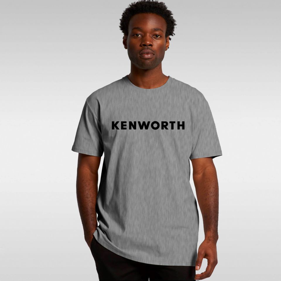 A man stands facing forward, wearing the SHIRT KW GREY WORD OUTLINE—a minimalist tee with bold black lettering across the chest. The light gray gradient background highlights this effortlessly stylish everyday shirt.