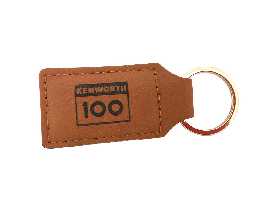 The KEYRING KW 100 YEARS TAN is a limited-edition tan leather keyring with visible stitching, metal key ring, and "KENWORTH 100" printed in black inside a rectangular border.