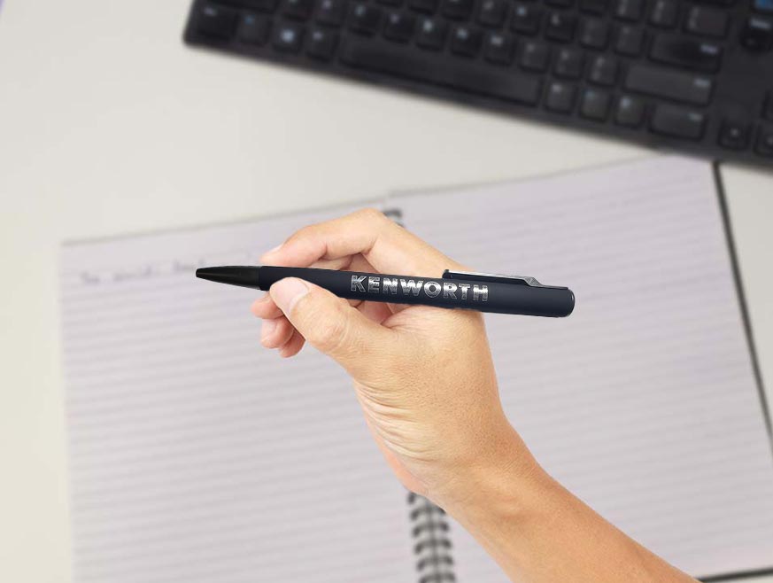 A hand holding a PEN KW BLACK recycled pen writes in a lined notebook, with a computer keyboard in the background—highlighting sustainable office supplies.