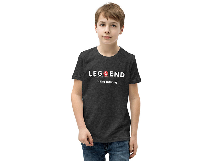 A young boy with short brown hair wears a TEE SHIRT KW KIDS LEGEND, a dark grey tee featuring "LEGEND in the making" with a red D20 die as the O, standing against a plain white background.
