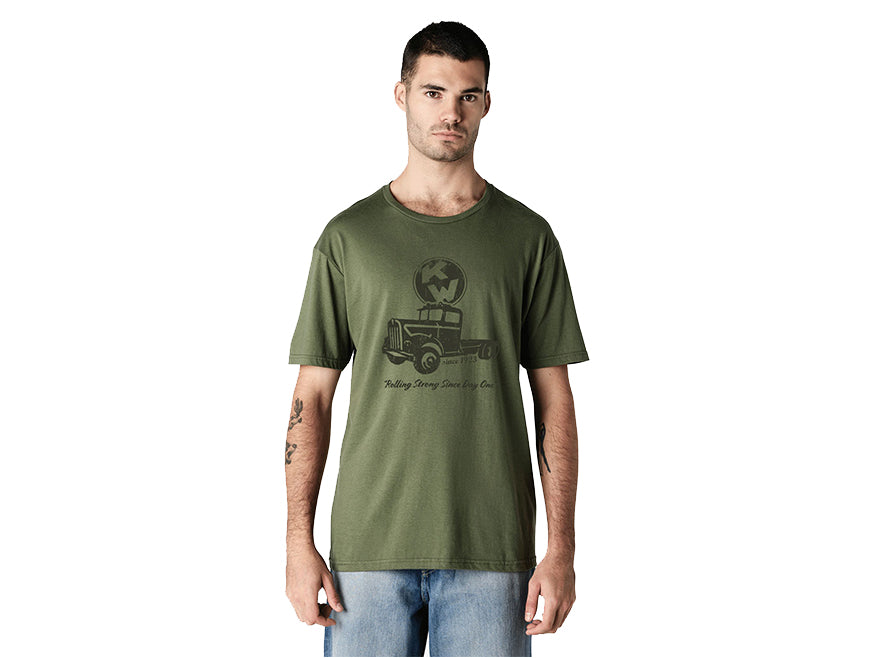 A man with short dark hair and light skin stands against a white background, wearing blue jeans and the TEE SHIRT KW ROLLING in military green, featuring a truck graphic and "Rolling Strong Since Day One" text. He looks at the camera.
