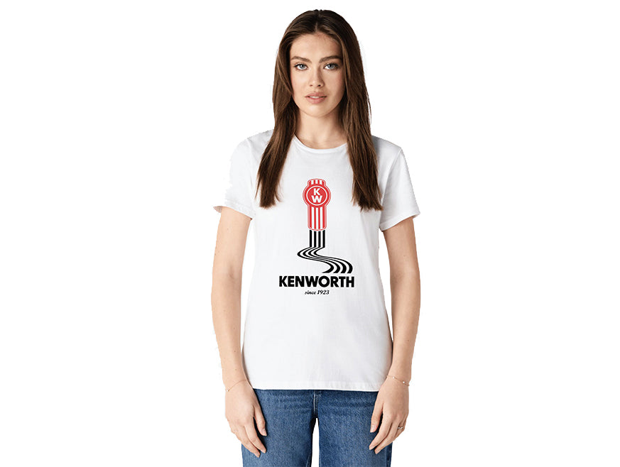 A woman with long brown hair wears the TEE SHIRT KW LINES, a white Kenworth tee featuring a red logo and winding black road graphic with subtle tyre marks. She stands facing forward against a plain white background.
