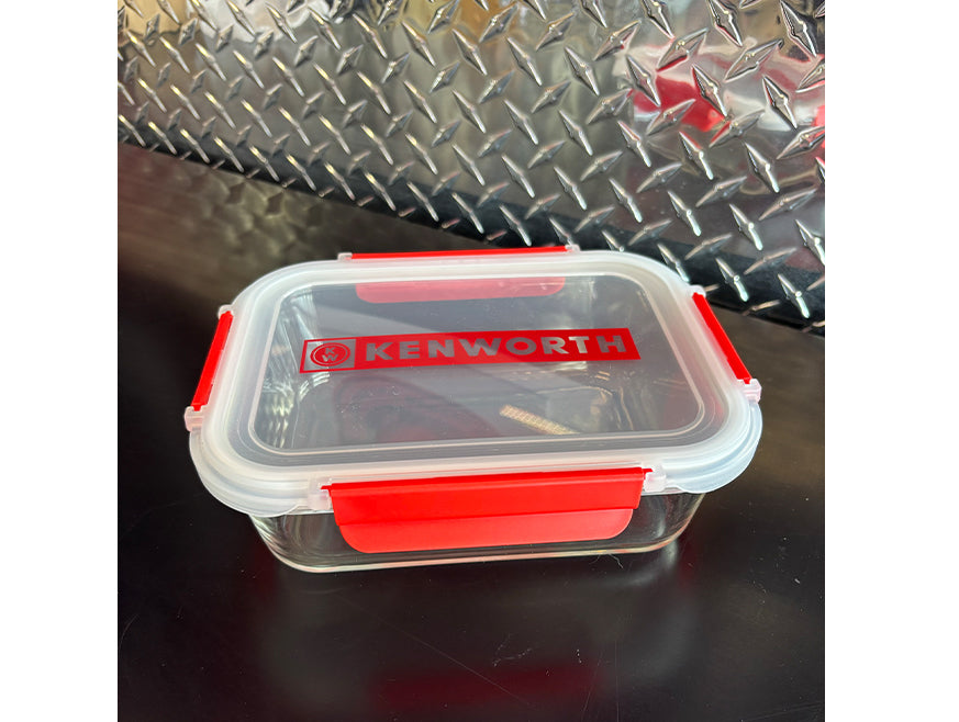 A clear rectangular borosilicate glass LUNCHBOX KW with red clips and a KENWORTH-labeled lid rests on a black surface in front of a metallic diamond plate background.