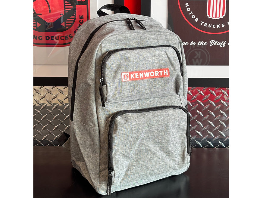 The BACKPACK KW GREY, a durable gray Kenworth backpack with black zippers and a red and white logo on the front pocket, rests on a black surface with framed automotive posters in the background.