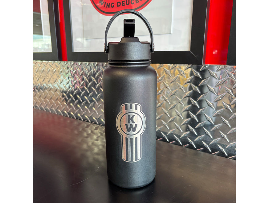 The BOTTLE KW VACUUM S/S, a black stainless steel bottle with a silver KW logo and straw lid, stands on a dark counter before a metallic diamond plate wall and red-and-black framed sign, ready to support your hydration needs.