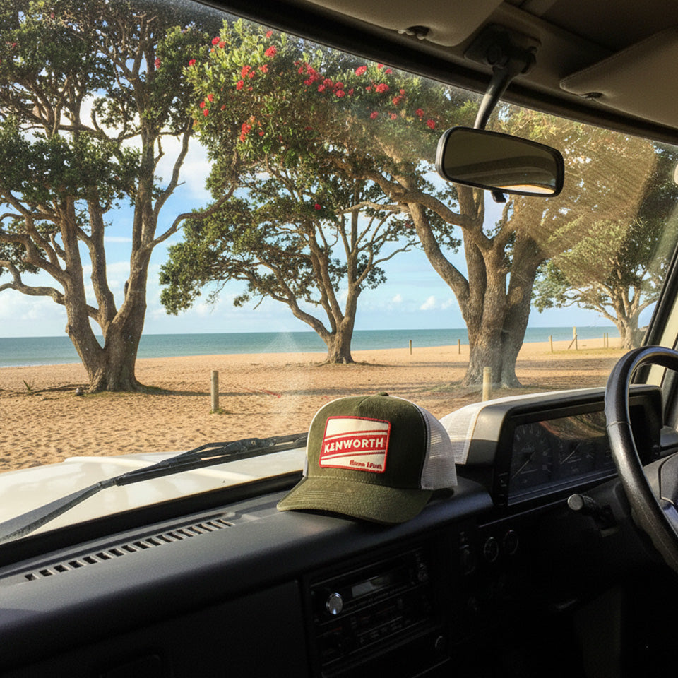 A Retro Kenworth Corduroy Cap with a green front and mesh back sits on a vehicle dashboard at a sandy beach, with trees, the ocean, and blue sky seen through the windshield.