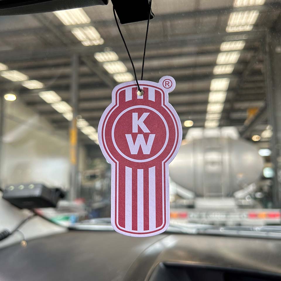 An AIR FRESHENER KW in red and white with an almond scent hangs from a car’s rearview mirror, set against a blurred industrial backdrop of large vehicles and bright overhead lights.