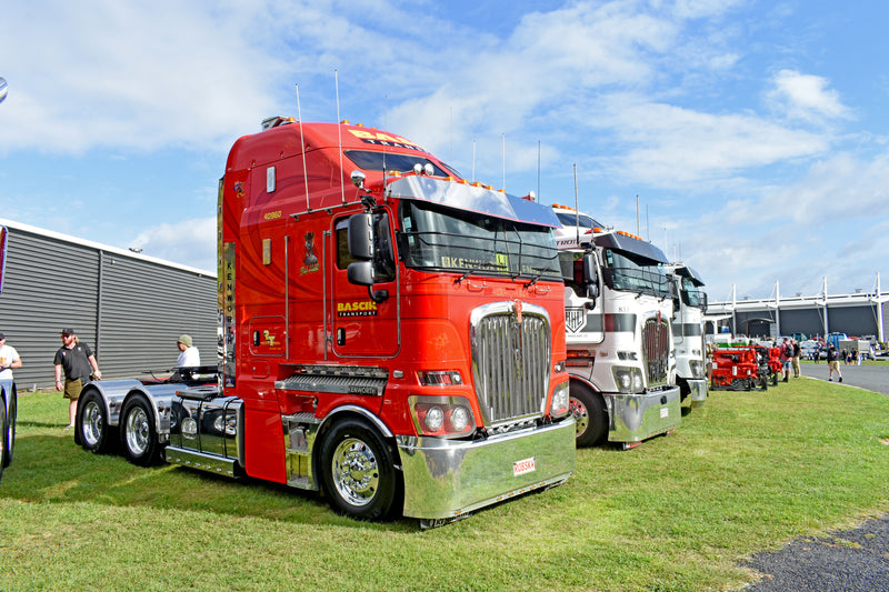 A row of large, brightly colored semi-trucks is parked on grass beside a building under a blue, partly cloudy sky. People stand nearby, admiring the trucks, with the closest one painted red.