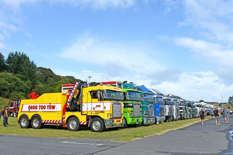 A row of colorful tow trucks and semi-trucks parked on grass beside a road under a partly cloudy sky, with people walking nearby and trees in the background.