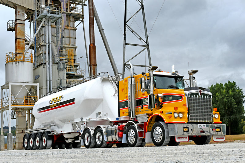 A yellow and white semi-truck with a large tanker trailer is parked on a gravel surface near an industrial facility with metal structures and silos. Trees and cloudy sky are in the background.