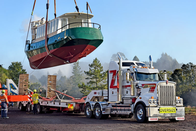 A large green and white boat is being lifted by a crane onto a flatbed trailer attached to a white semi-truck with an OVERSIZE sign. Workers in safety gear guide the operation outdoors near trees.