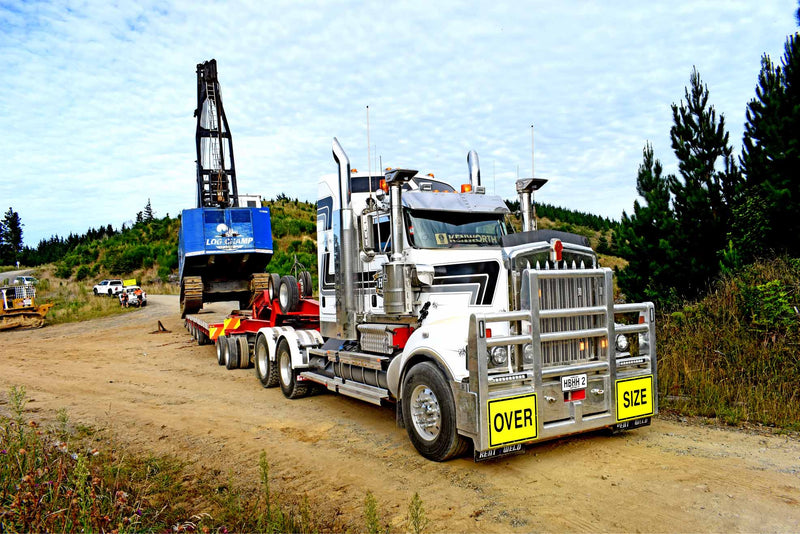 A large silver semi-truck with OVER SIZE signs hauls a blue excavator on a flatbed trailer along a dirt road, with trees and grassy hills in the background under a partly cloudy sky.