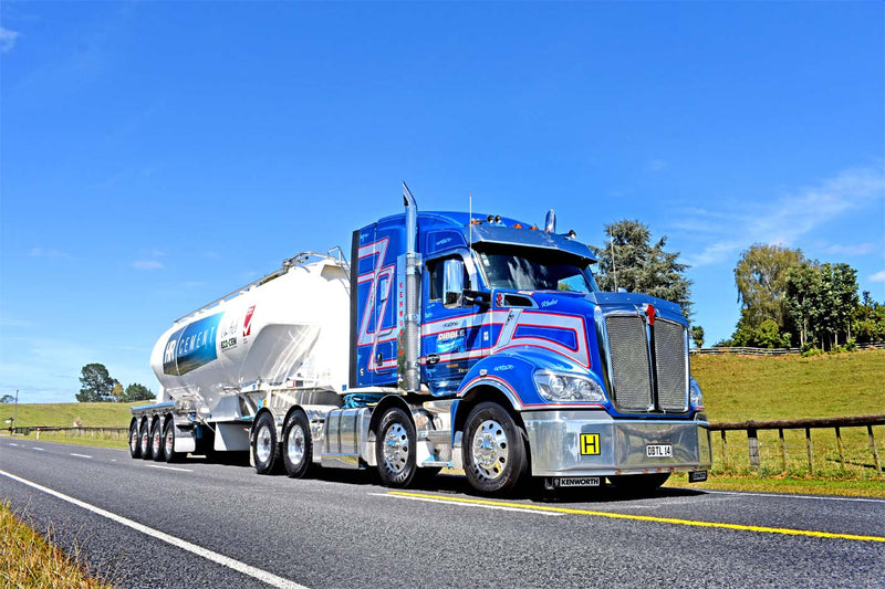 A blue semi-truck with red and white stripes is hauling a white tanker trailer on a rural road with green fields, trees, and a clear blue sky in the background.